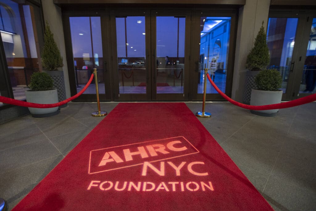A red carpet featuring the AHRC NYC Foundation logo leads to a set of glass double doors behind velvet rope stanchions.