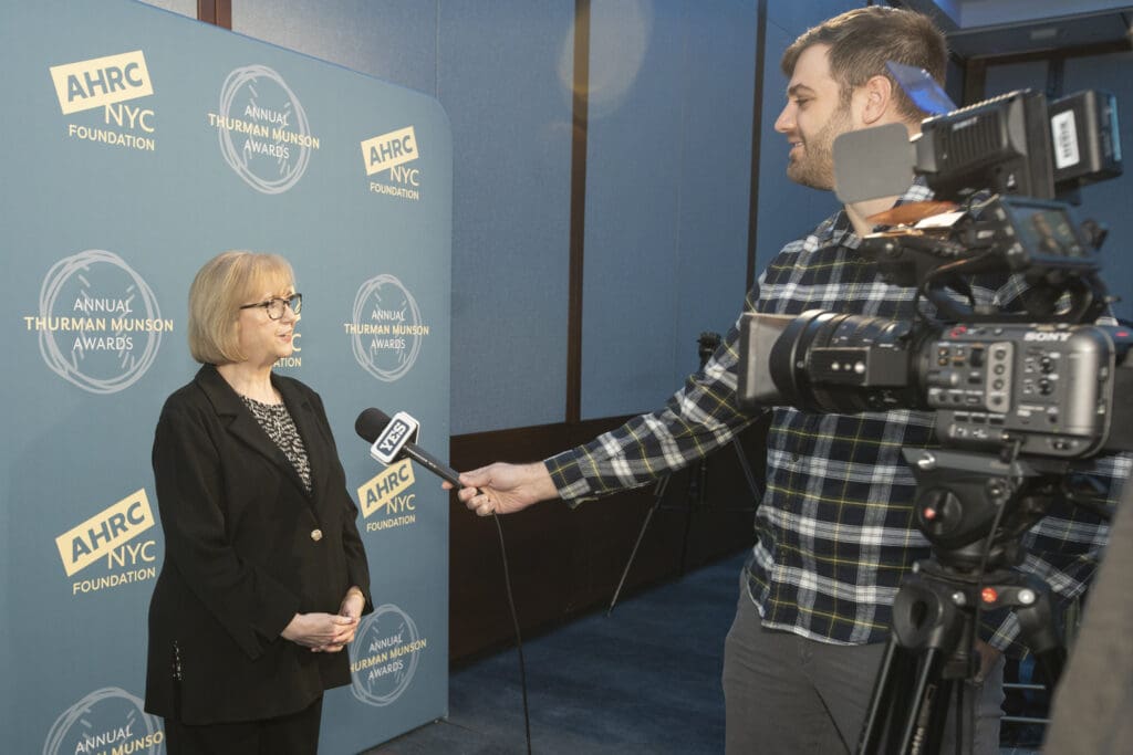 A woman in a black blazer being interviewed by a person holding a Yes Network microphone in front of an AHRC NYC Foundation backdrop.