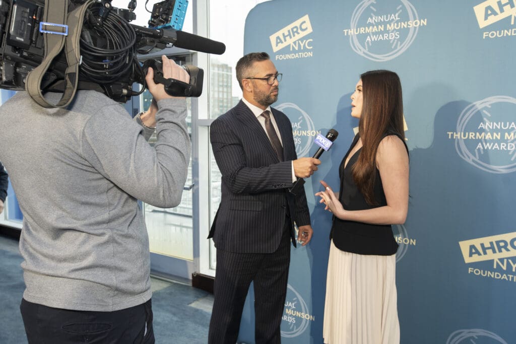 A reporter in a suit interviews a woman holding a microphone in front of an AHRC NYC Foundation backdrop.