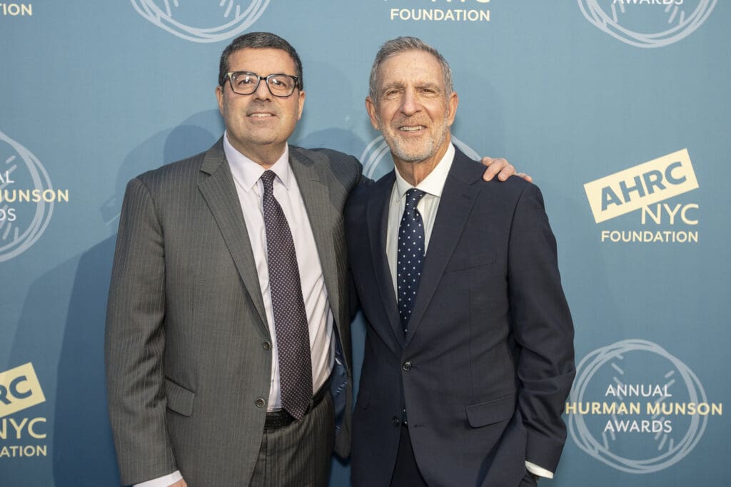 Two men in suits posing together in front of an AHRC NYC Foundation event backdrop.