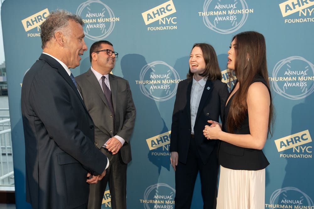 Two men in suits and two women in business attire talking in front of an AHRC NYC Foundation backdrop for the Annual Thurman Munson Awards.