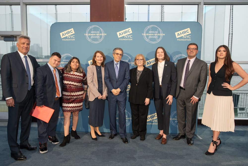 Nine people stand in a line on a stage in front of an AHRC NYC Foundation Annual Thurman Munson Awards backdrop.