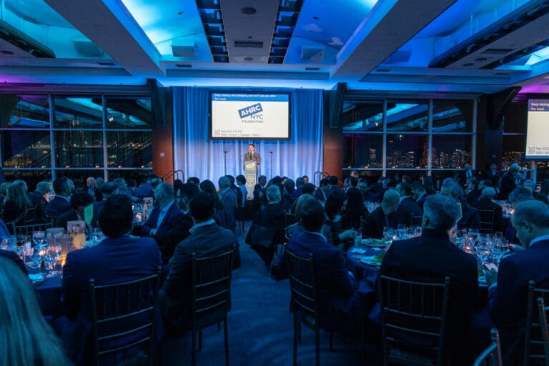 A man stands at a podium giving a presentation to a banquet hall of seated guests in front of a screen displaying the AHRC NYC Foundation logo.