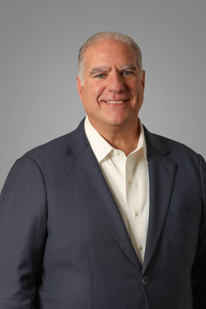 A middle-aged man with gray hair smiling, wearing a navy blue suit jacket and a white dress shirt.